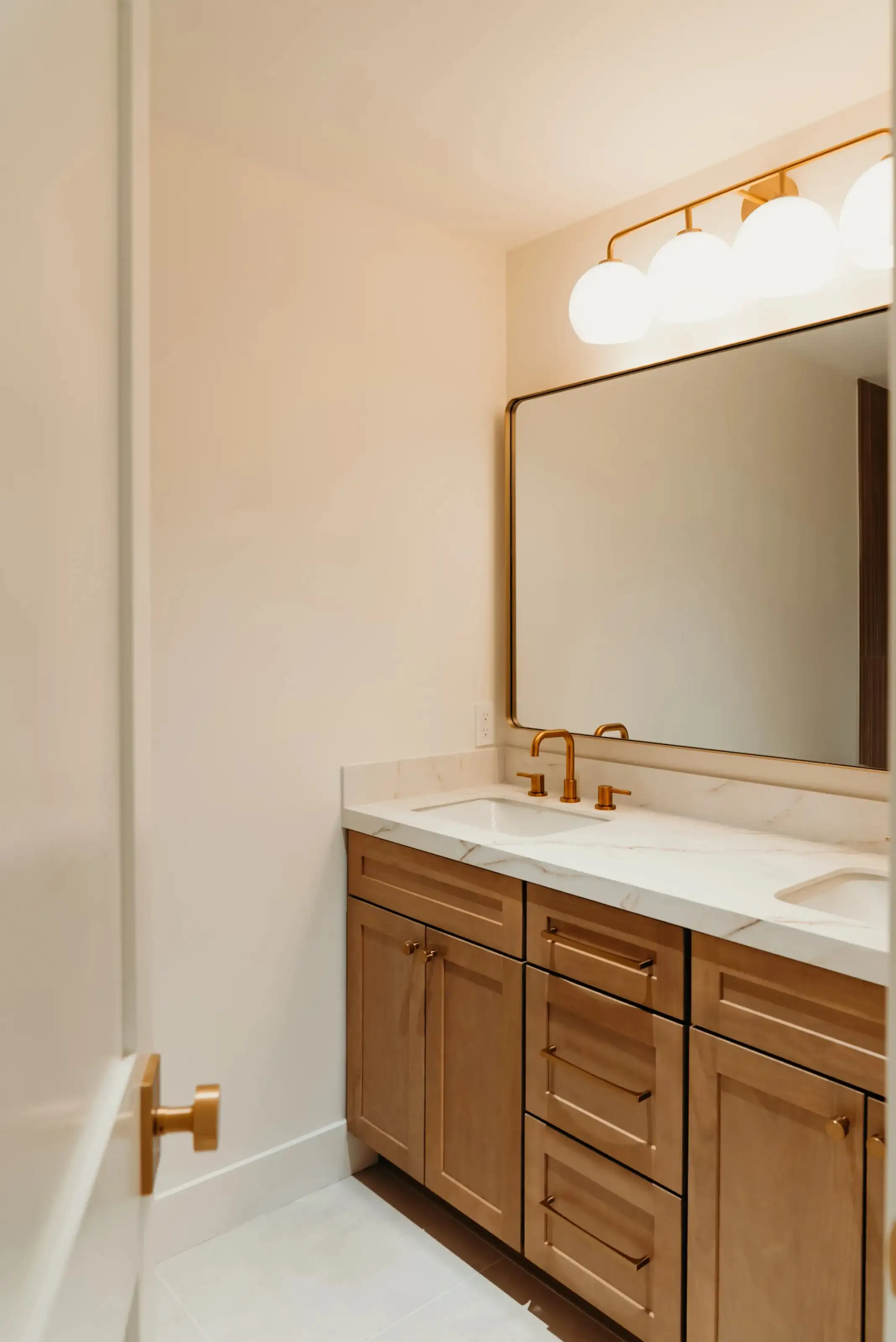 Bathroom vanity remodel with wood cabinets, white marble countertop, gold faucet, and large mirror with globe lights