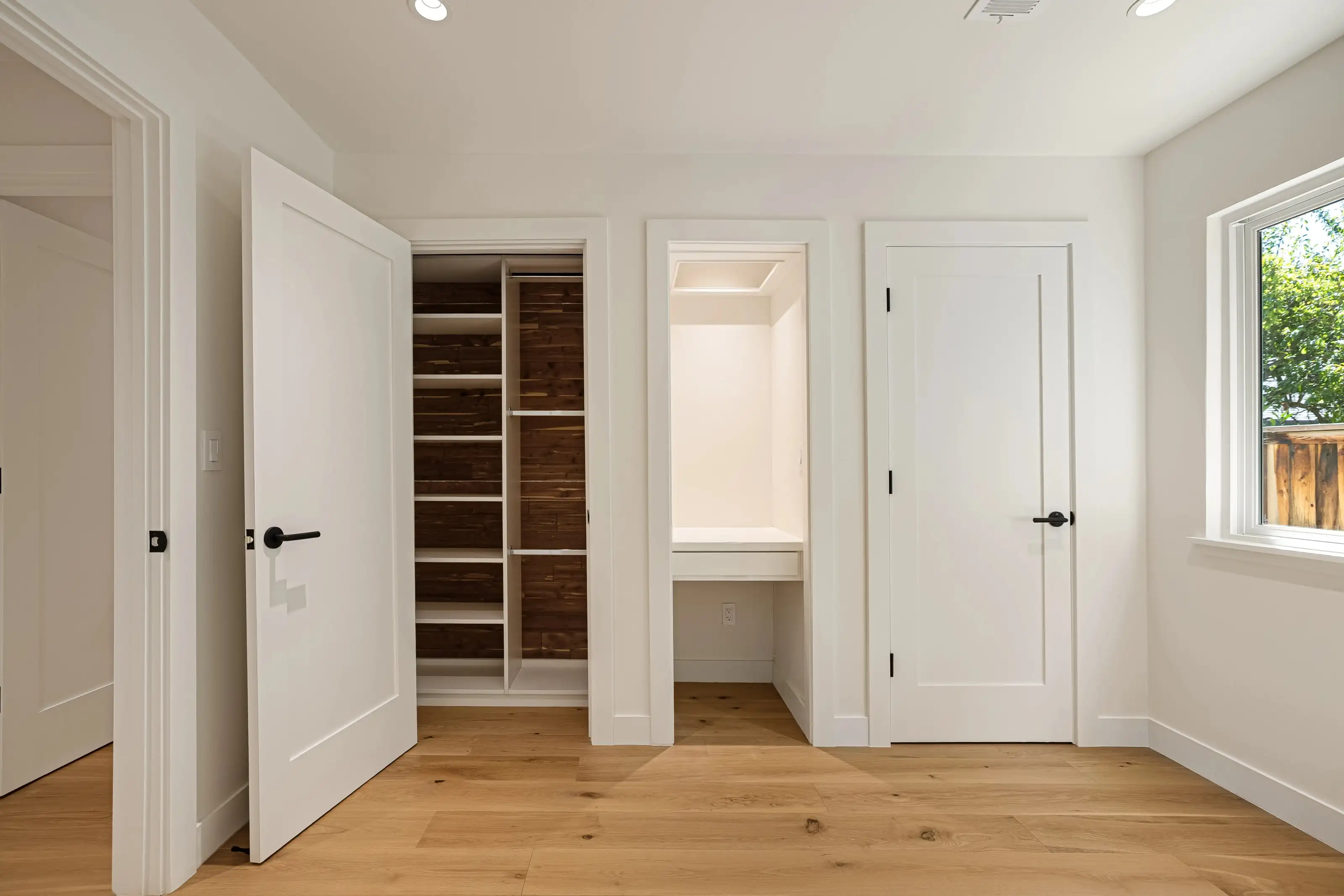 Open closet with dark wood shelving and a small built-in desk nook with lighting, white shaker doors, light hardwood floor