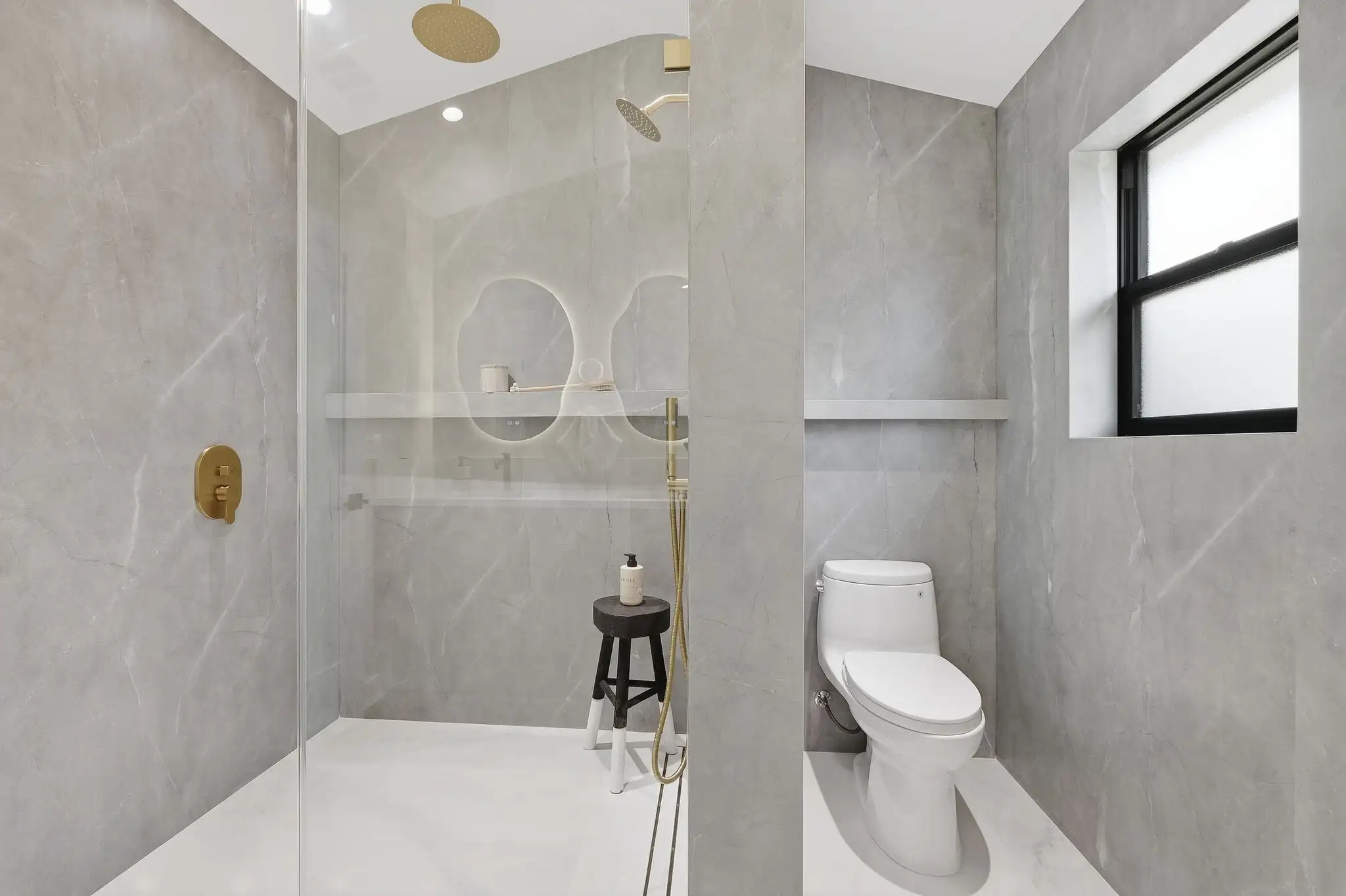 Modern bathroom remodel with gray marble tile walls, gold rain shower fixtures, glass partition, and white toilet in San