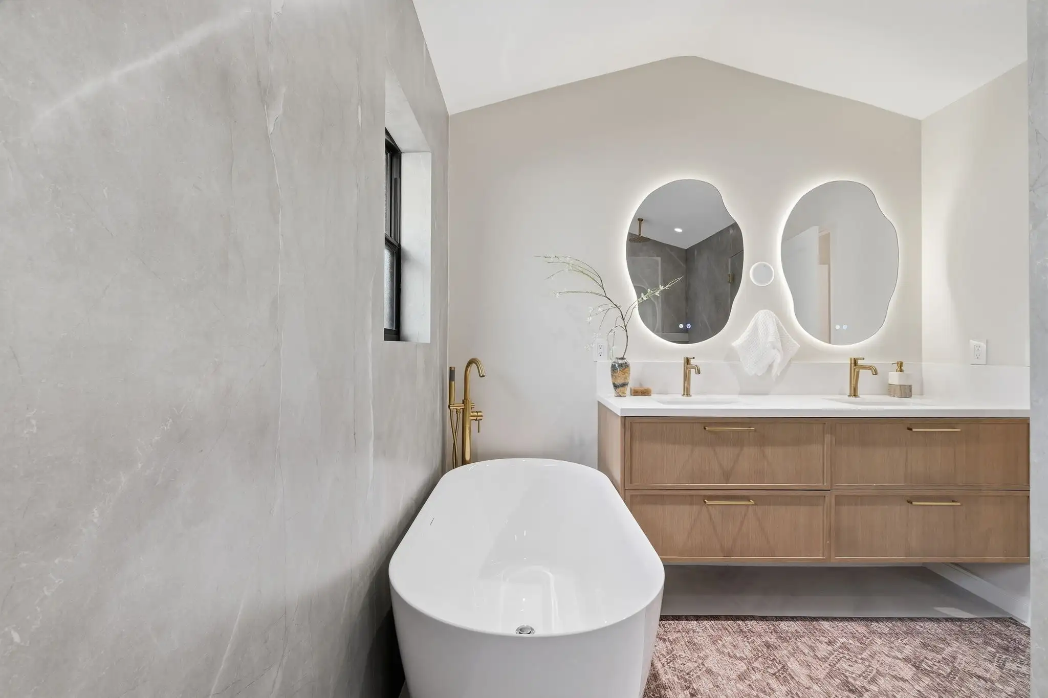 Freestanding white soaking tub with brass floor faucet next to a floating wood vanity with dual sinks and backlit mirrors